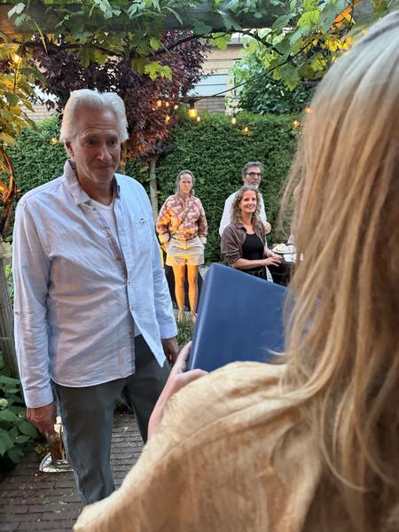 A group of people are gathered in a lush garden at Koetlaan, Vogelwijk, Den Haag, enjoying a relaxed evening under string lights
